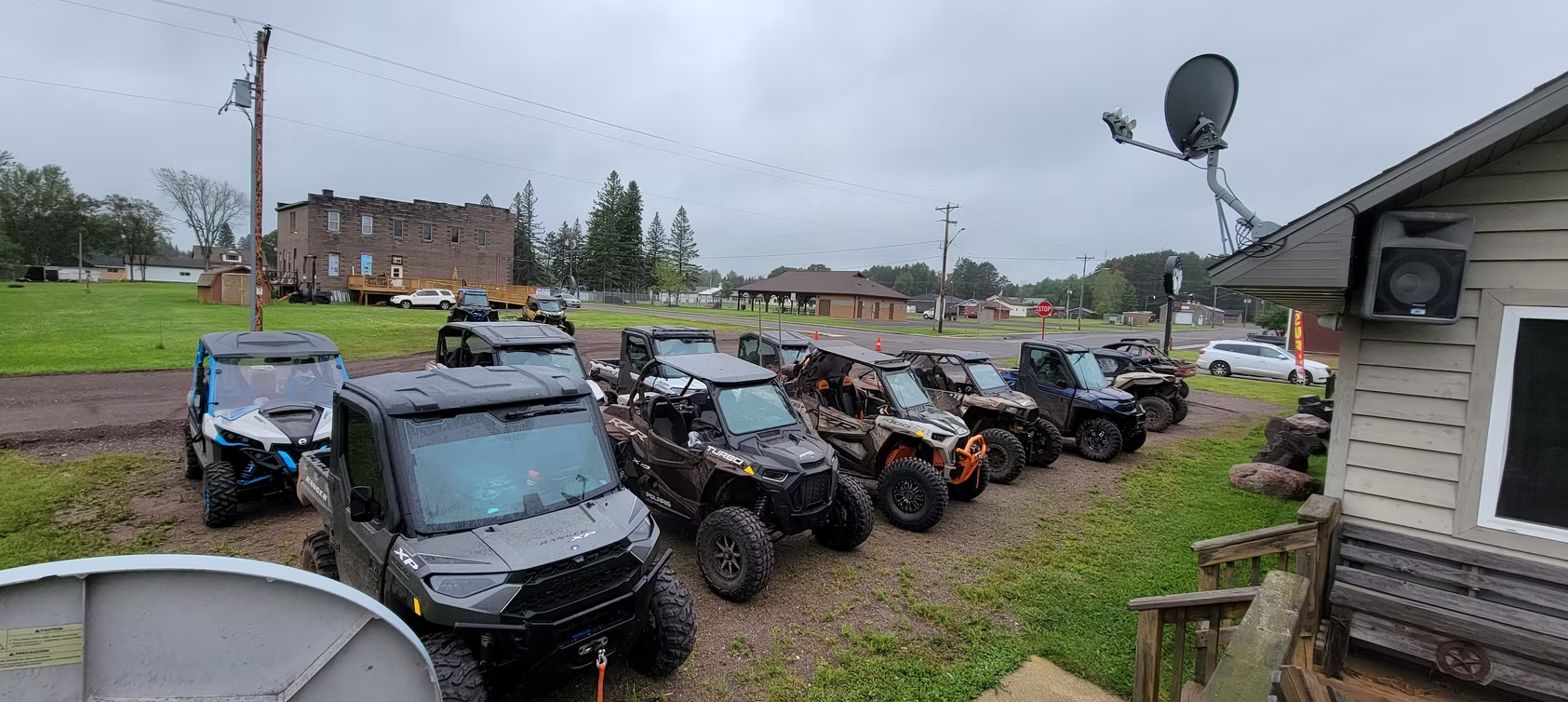 Row of damaged off-road vehicles parked in a muddy lot beside a house under cloudy skies