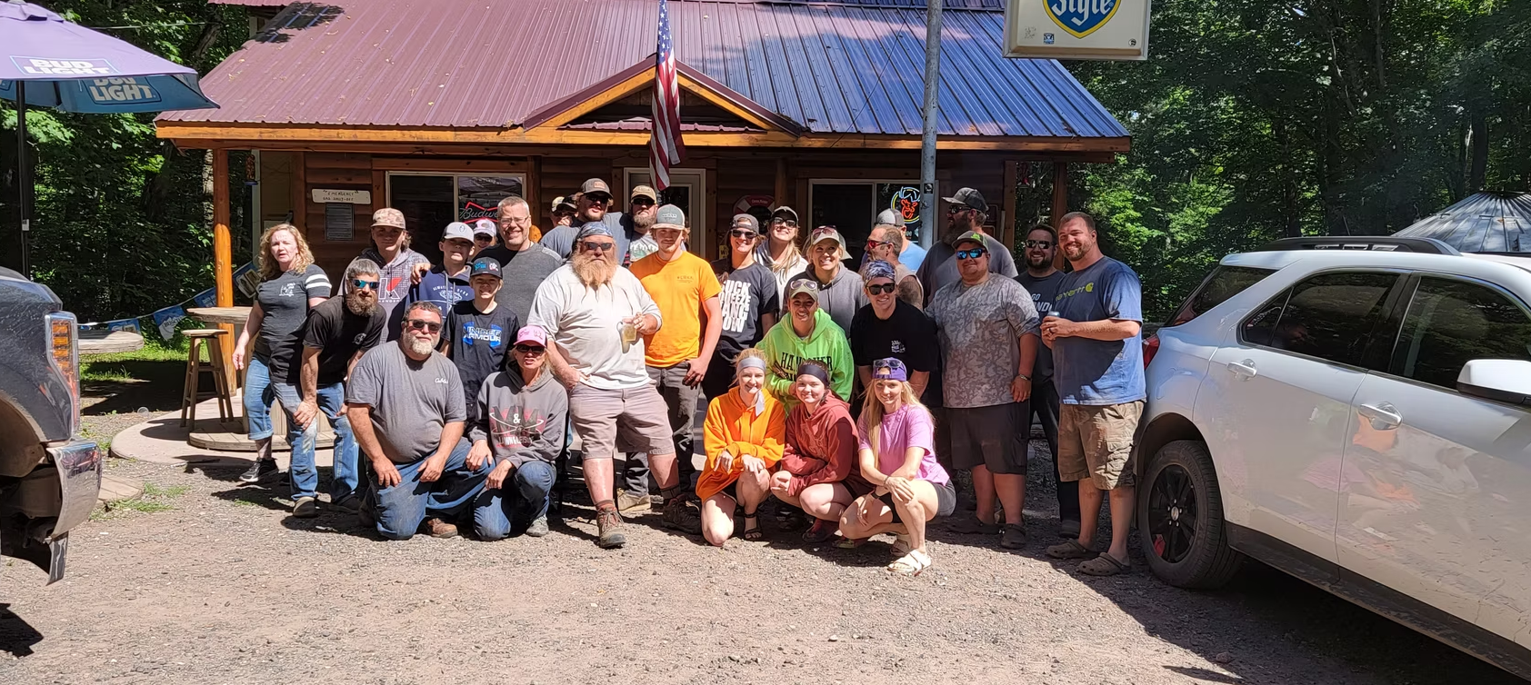 Large group posing outside a wooden building, with a white car parked beside them.