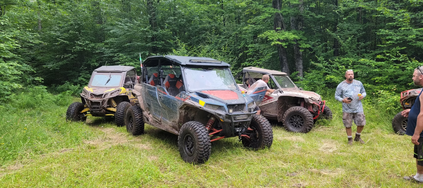 Three muddy off-road vehicles parked on a grassy trail in a wooded area, with people standing nearby.