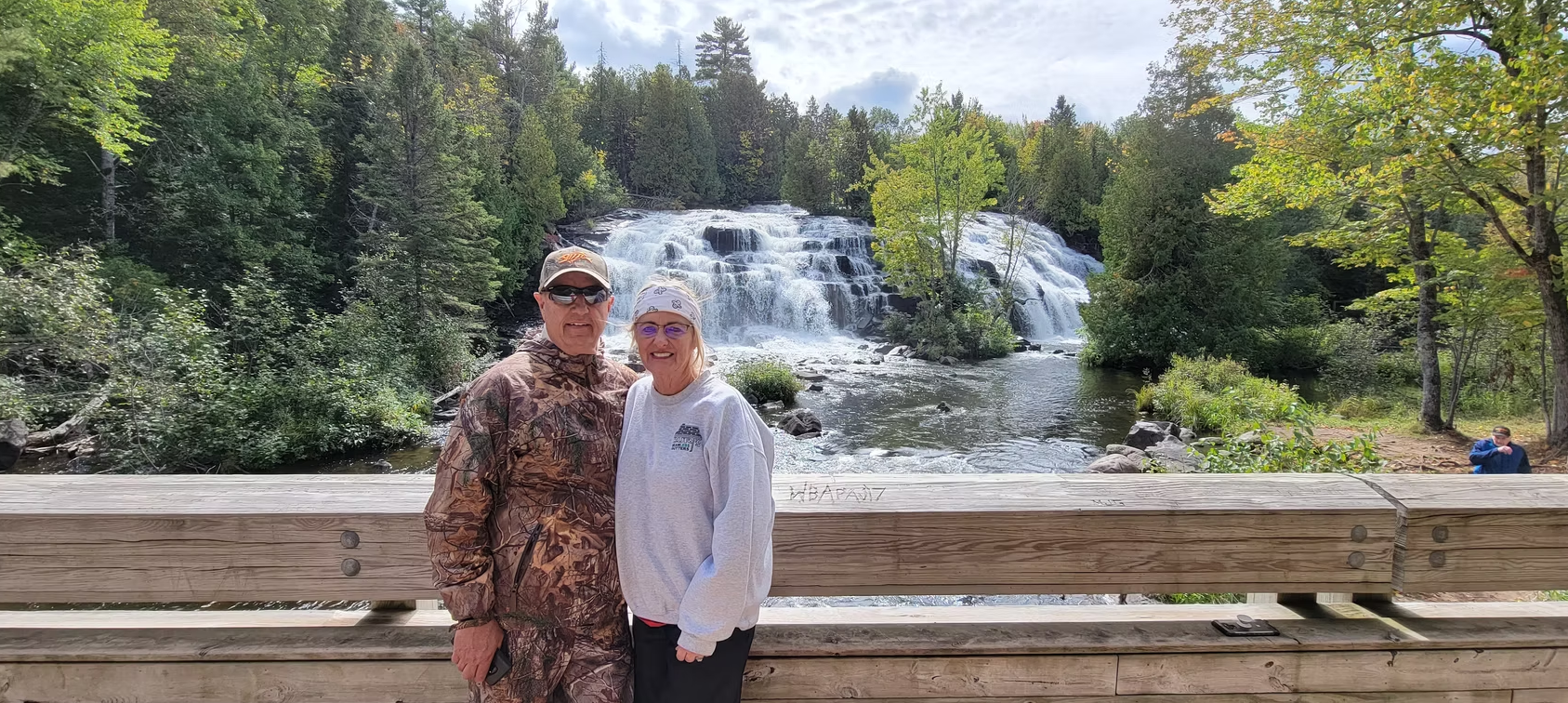 Two people posing by a railing in front of a multi-tiered waterfall and forested landscape