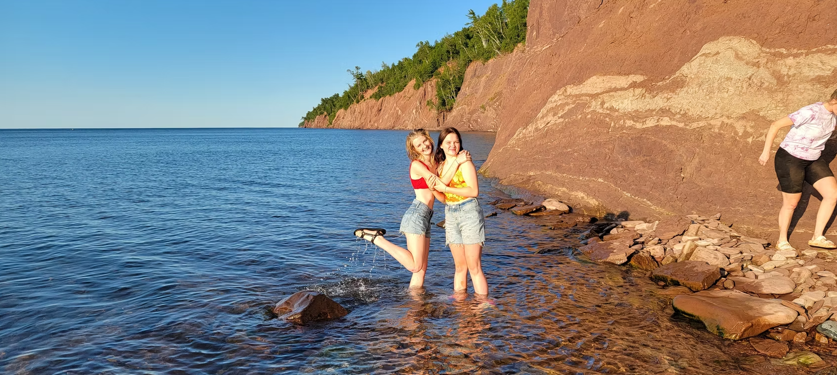 Two people standing on rocks in shallow ocean water beside a rocky shoreline