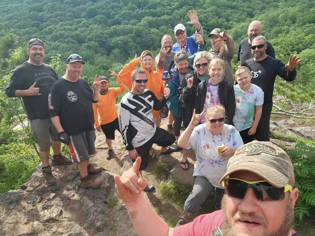 Group of hikers posing on a rocky overlook, smiling and waving in bright outdoor gear.