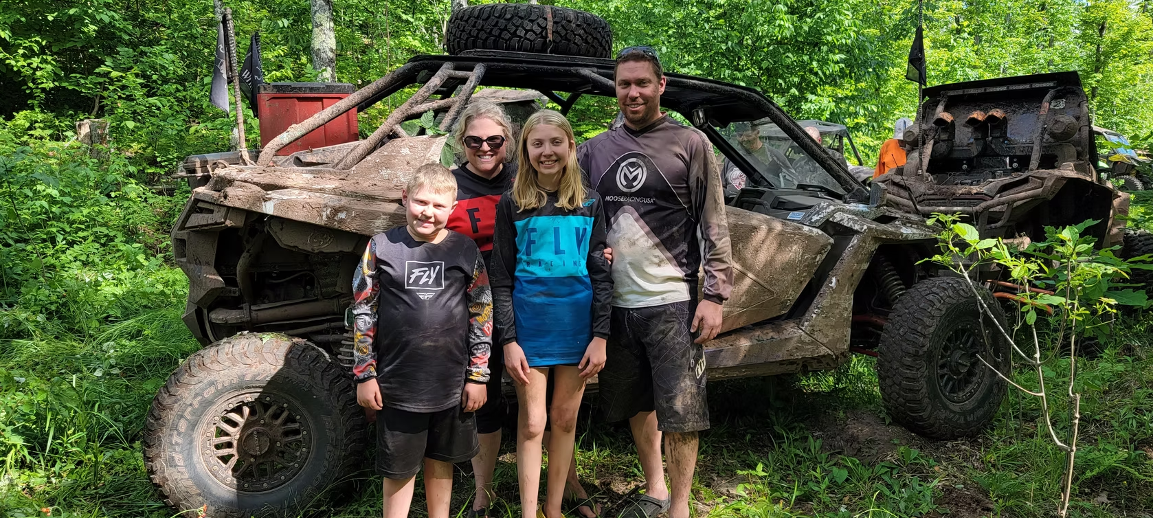 Three people stand beside a muddy off-road vehicle in a green wooded area.
