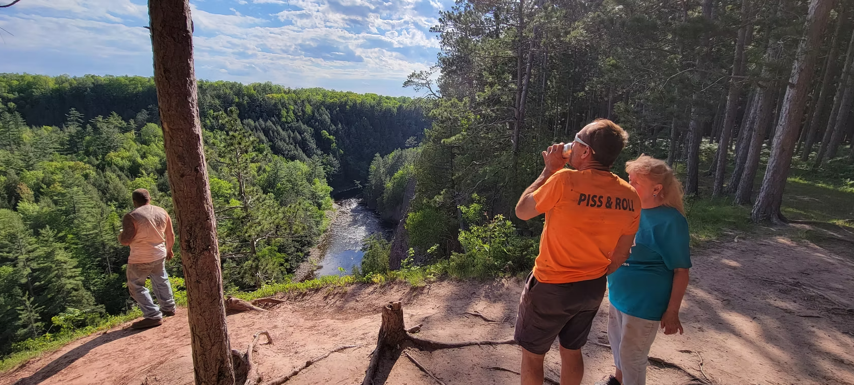 Three people overlooking a forested waterfall from a sunny overlook, one taking a photo.