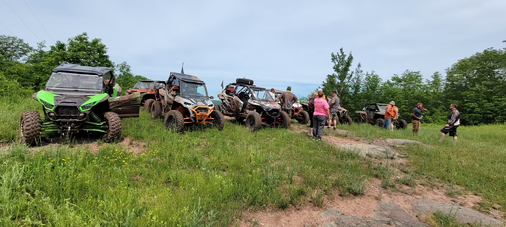 Mud-splattered off-road vehicles and a few people gathered on a grassy trail under a cloudy sky.