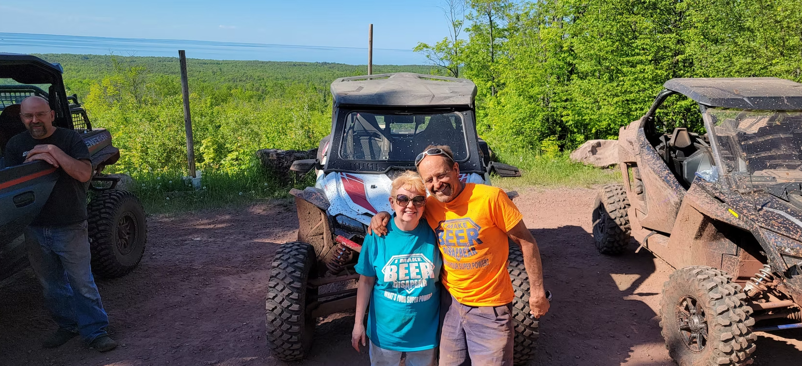 Two people posing in front of an off-road buggy on a sunny trail, with fields and another vehicle nearby.