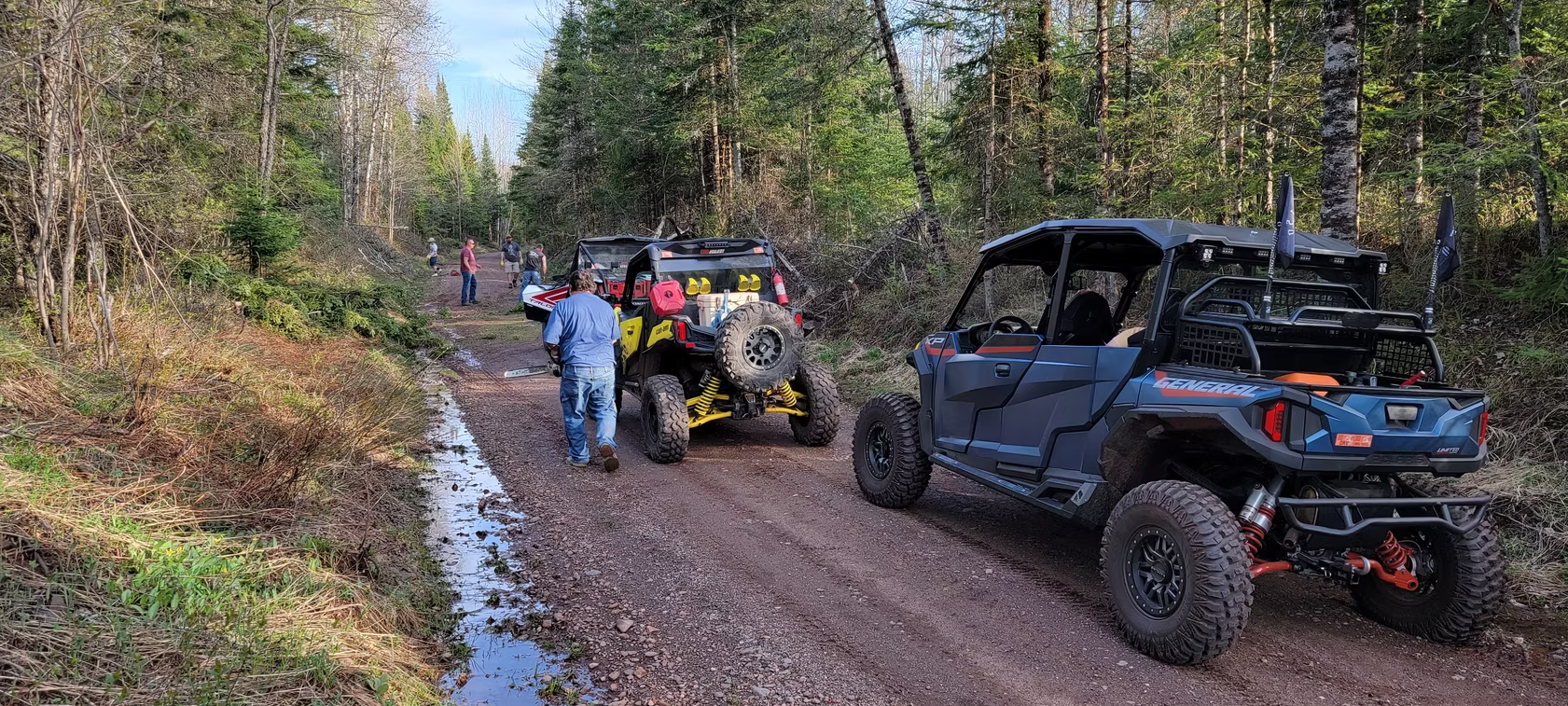 Off-road vehicles and people on a muddy forest trail, with a blue UTV in the foreground.