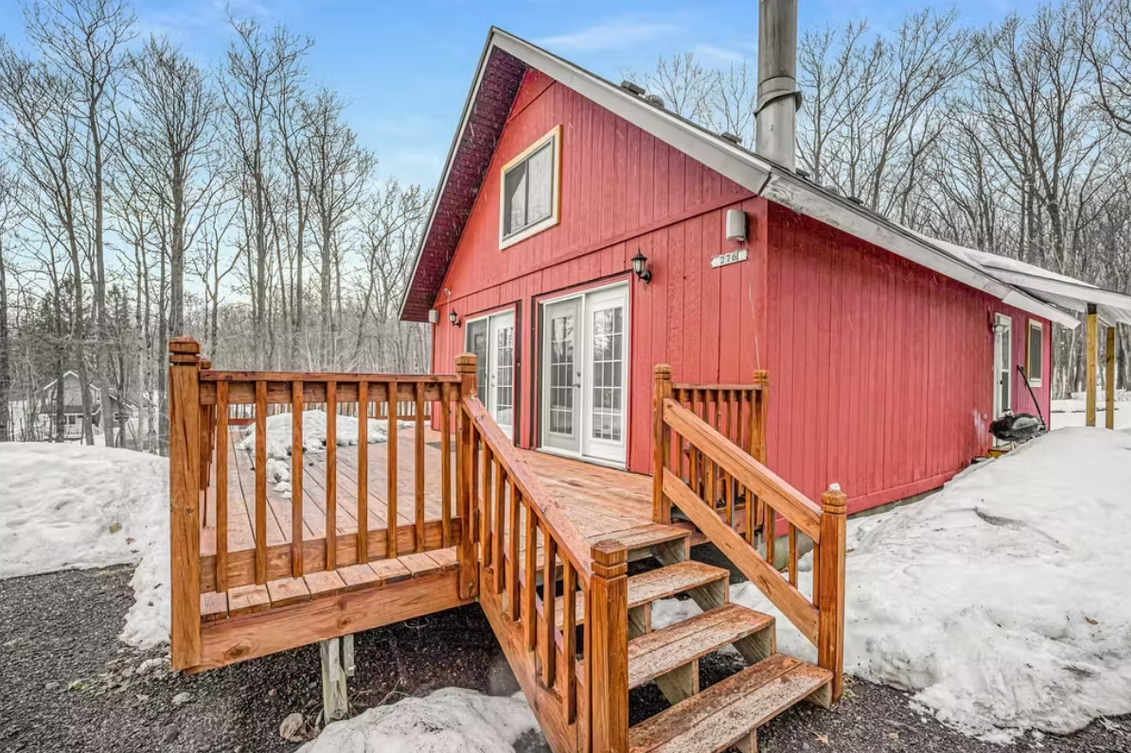 Red cabin with wooden deck and steps in a snowy wooded yard