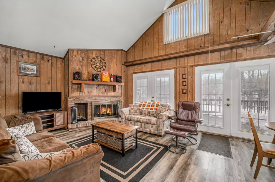 Wood-paneled living room with stone fireplace, brown sofas, TV, and French doors to a deck