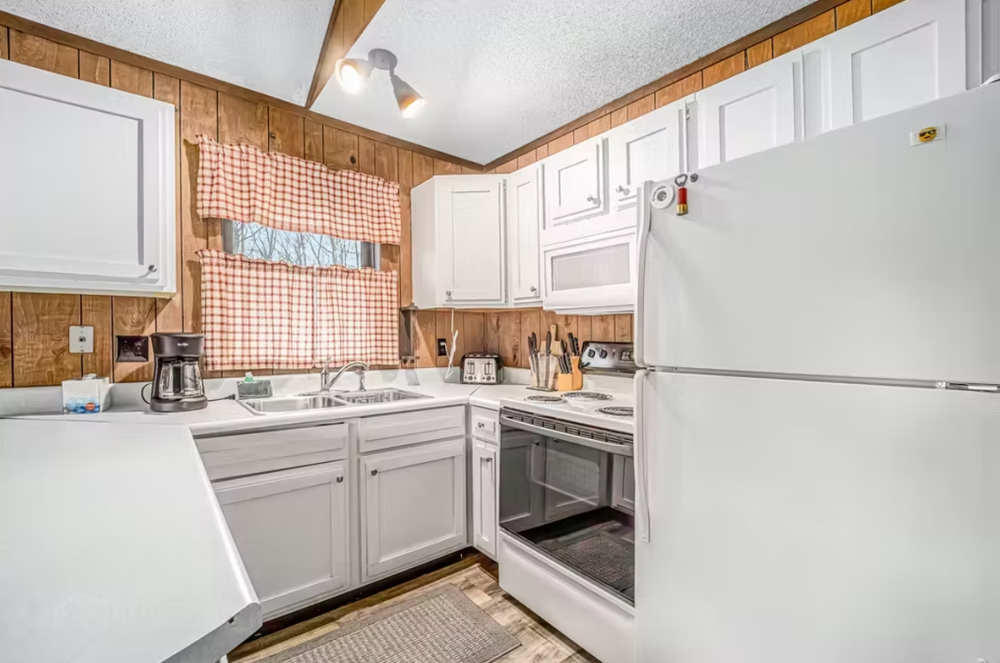 Bright white kitchen with cabinets, stove, refrigerator, and checkered curtains under vaulted wood ceiling