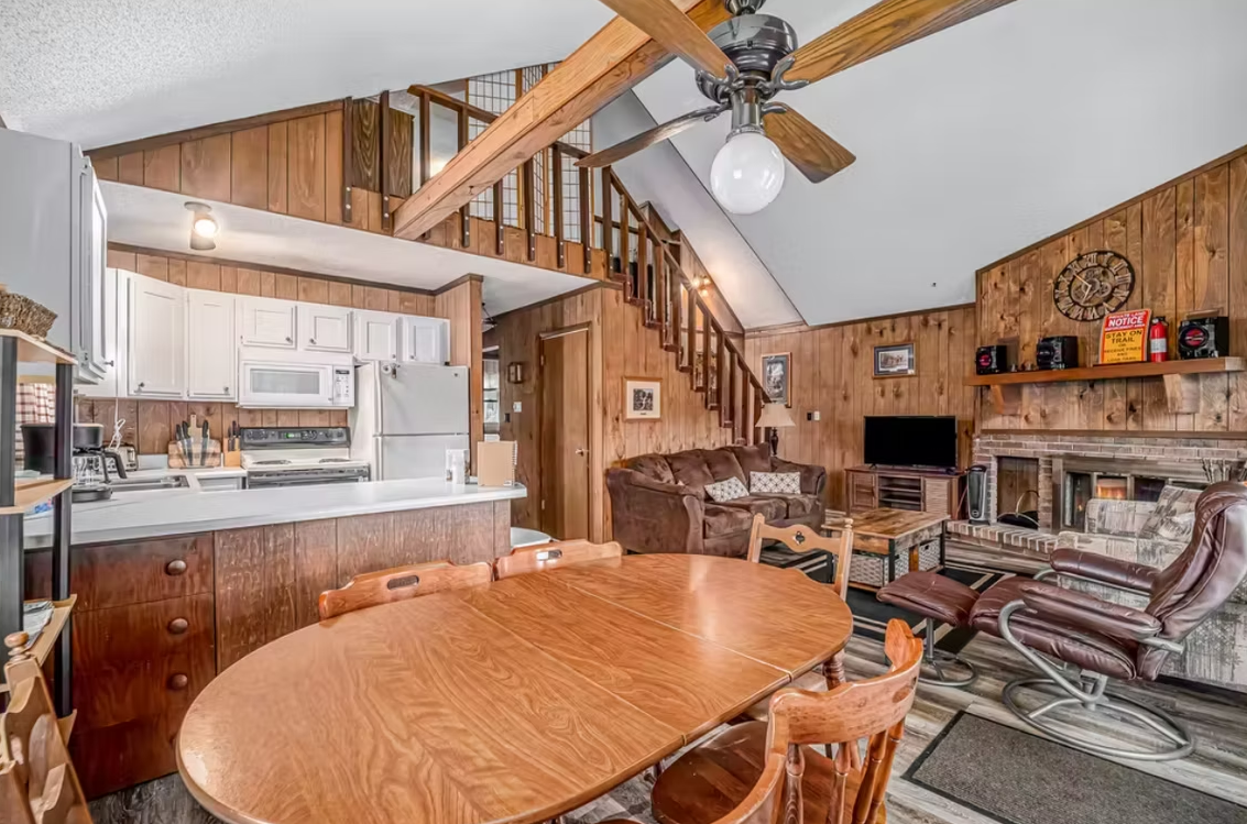 Wood-paneled cabin interior with dining table, kitchen pass-through, staircase, and fireplace