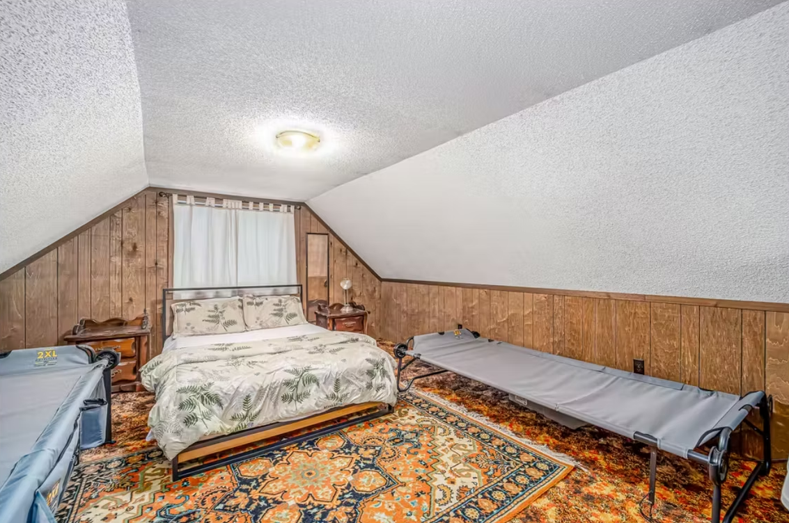Small attic bedroom with three beds, wood paneling, patterned rug, and a curtained window.
