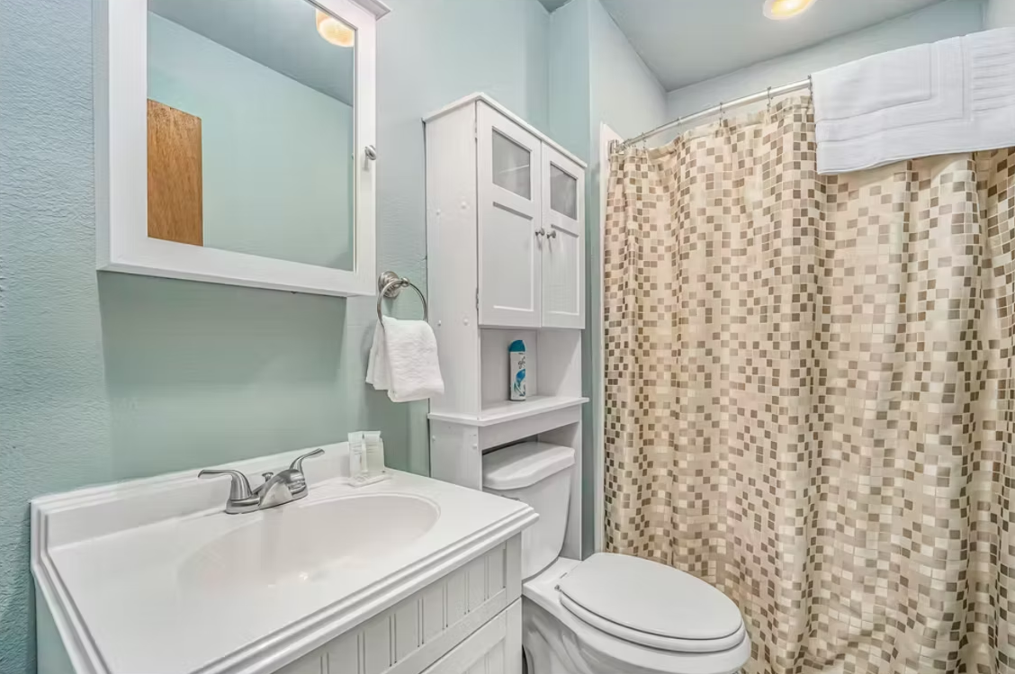 Small light-blue bathroom with white sink, toilet, cabinet, mirror, and patterned shower curtain.