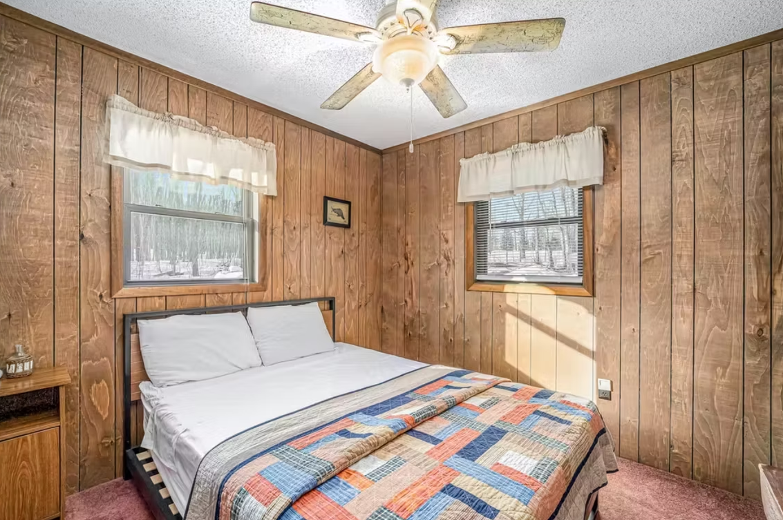 Cozy wood-paneled bedroom with a bed, two windows, and a ceiling fan