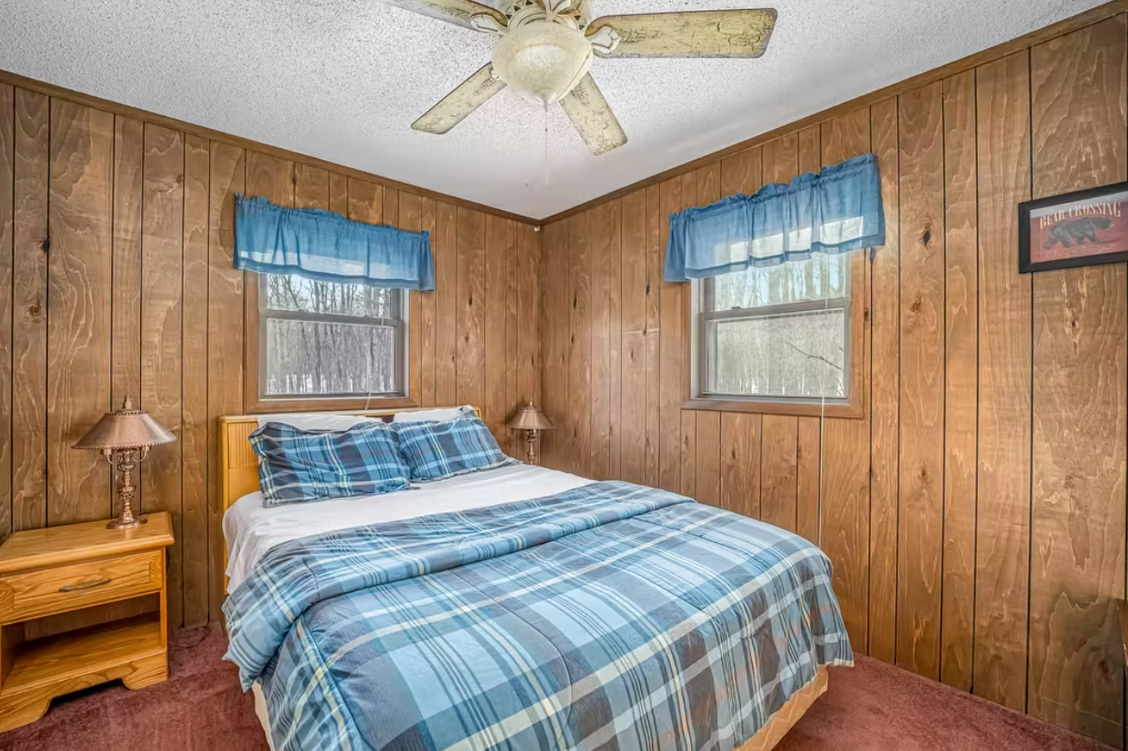 Small wood-paneled bedroom with blue plaid bedding, two windows, and a ceiling fan