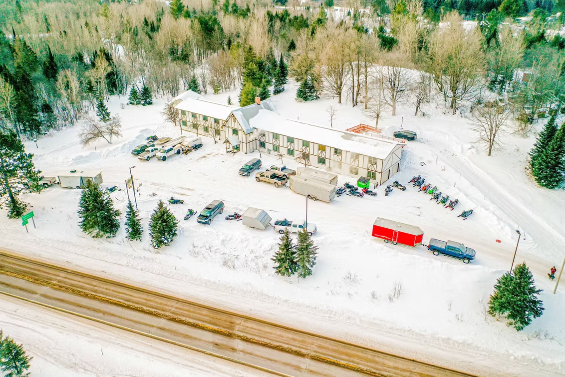 Snow-covered roadside building with parked vehicles beside a forest and railway tracks.