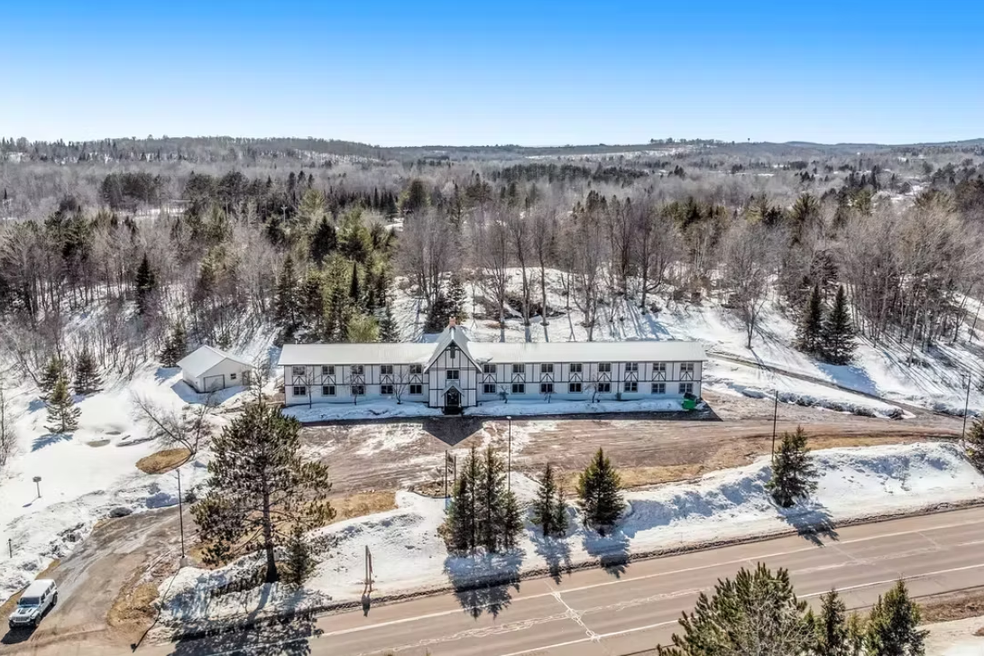 Aerial view of a snow-covered building beside a road, surrounded by trees and winter landscape.
