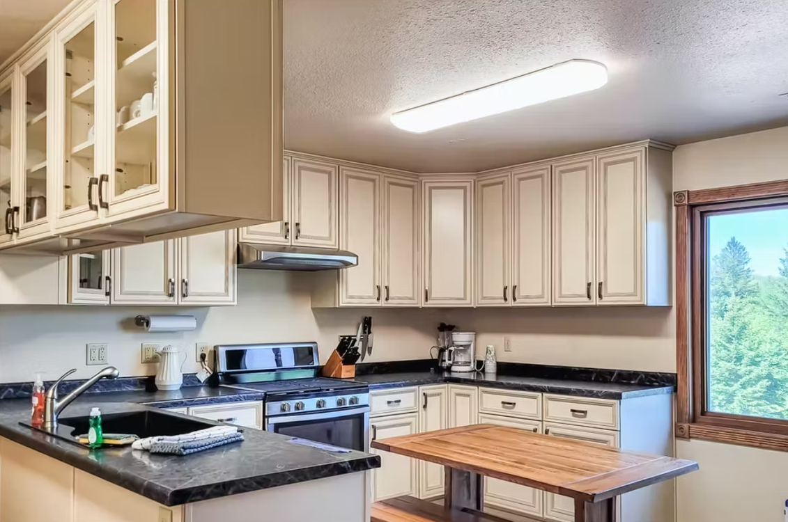 Bright kitchen with cream cabinets, black countertops, a stove, island, and large window with trees outside