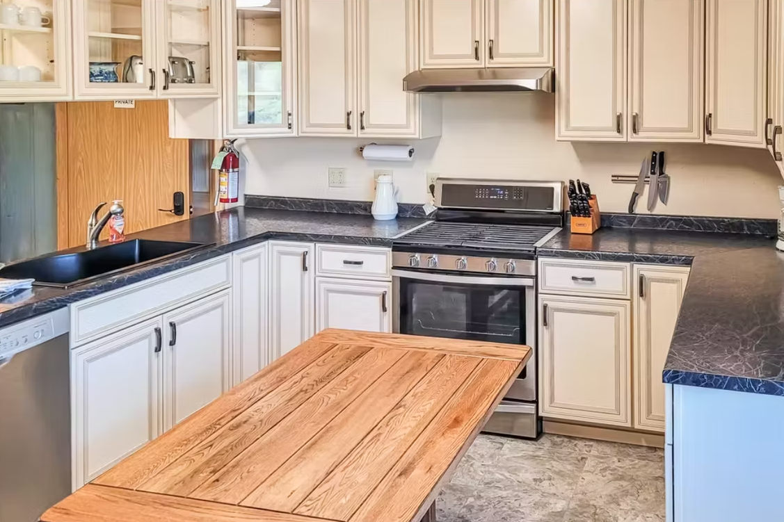 Bright kitchen with cream cabinets, black countertops, stainless stove, and a wooden island.