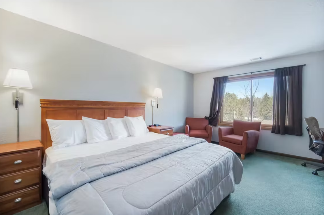 Bright hotel bedroom with king bed, teal carpet, armchairs, and a large window with curtains