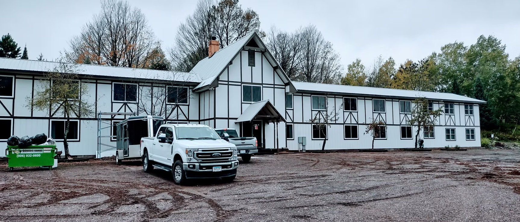 White two-story building with black trim and parked cars on a muddy lot under an overcast sky