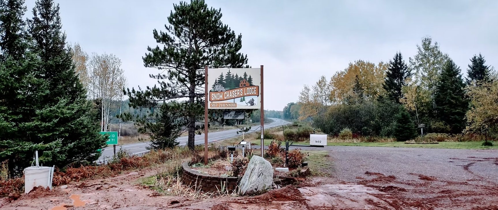 Rustic roadside sign in a wooded area with a gravel clearing and evergreen trees.