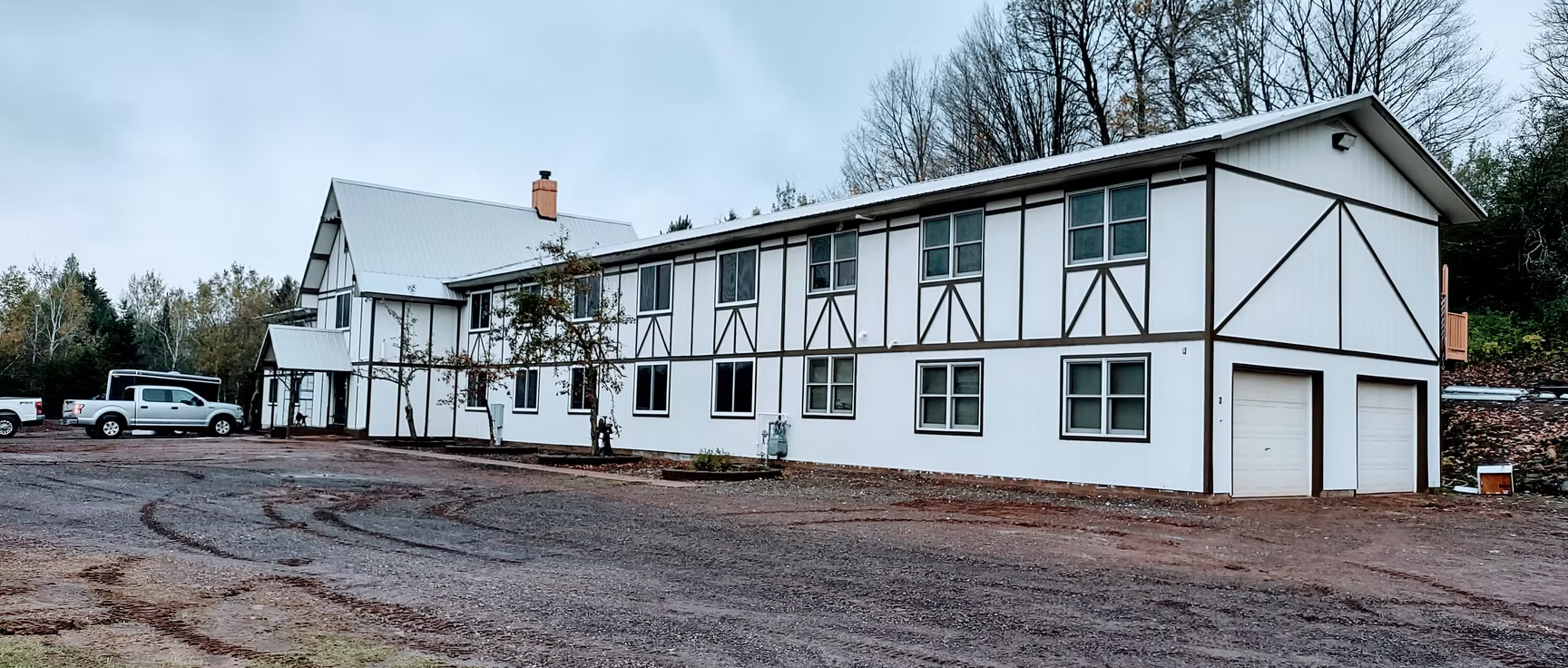 White two-story half-timbered building with a garage, seen across a muddy lot on a cloudy day