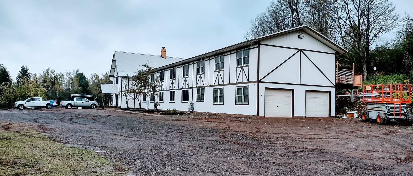 White two-story building with garage doors in a muddy lot, trees and utility trucks in the background