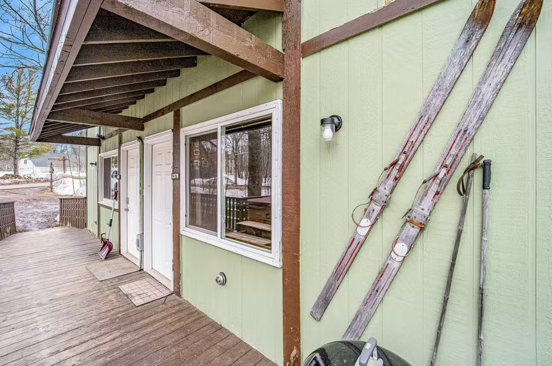 Exterior deck beside a pale green house with windows, wood posts, and skis mounted on the wall