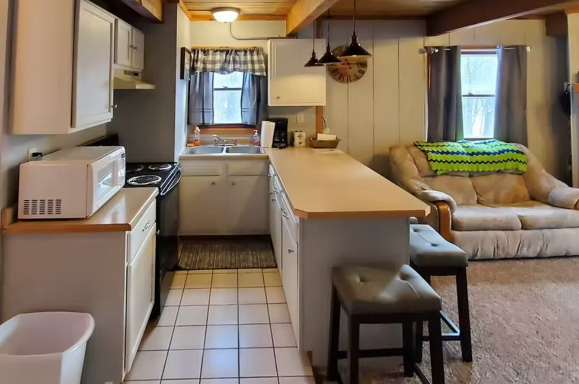 Compact kitchen opening to a small living area with beige walls, white appliances, and a window seat.