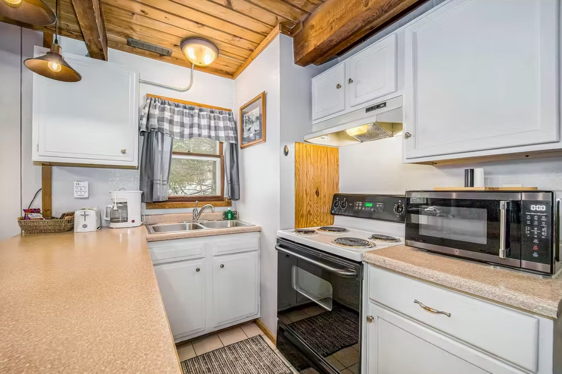 Compact kitchen with white cabinets, wood countertops, stove, microwave, and sink under a window