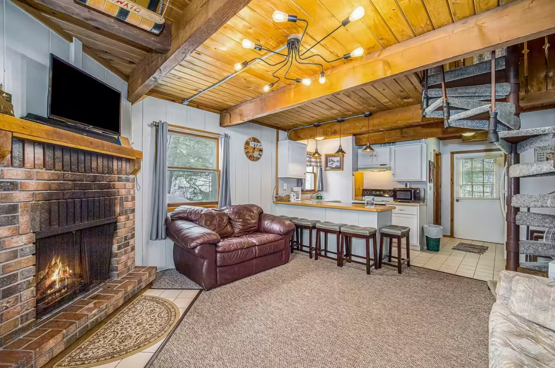 Cozy rustic living room with stone fireplace, leather sofa, and open kitchen with wood-beamed ceiling