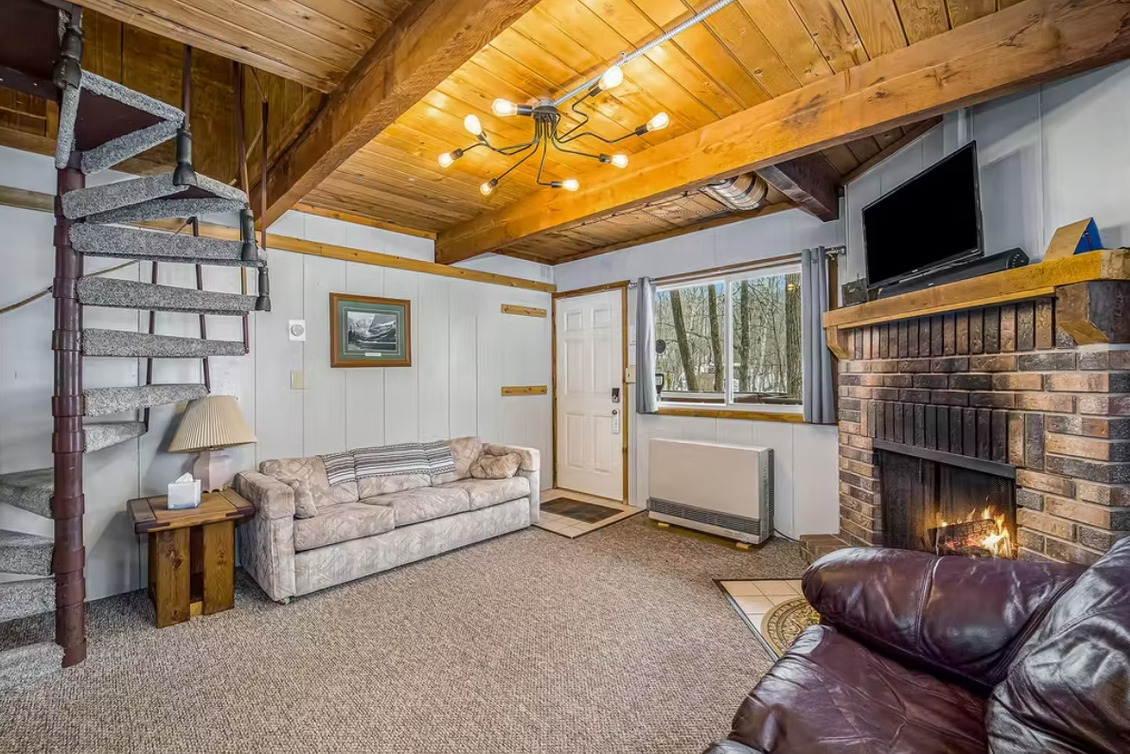 Cozy rustic living room with wood ceiling, stone fireplace, sofa, spiral stairs, and large window