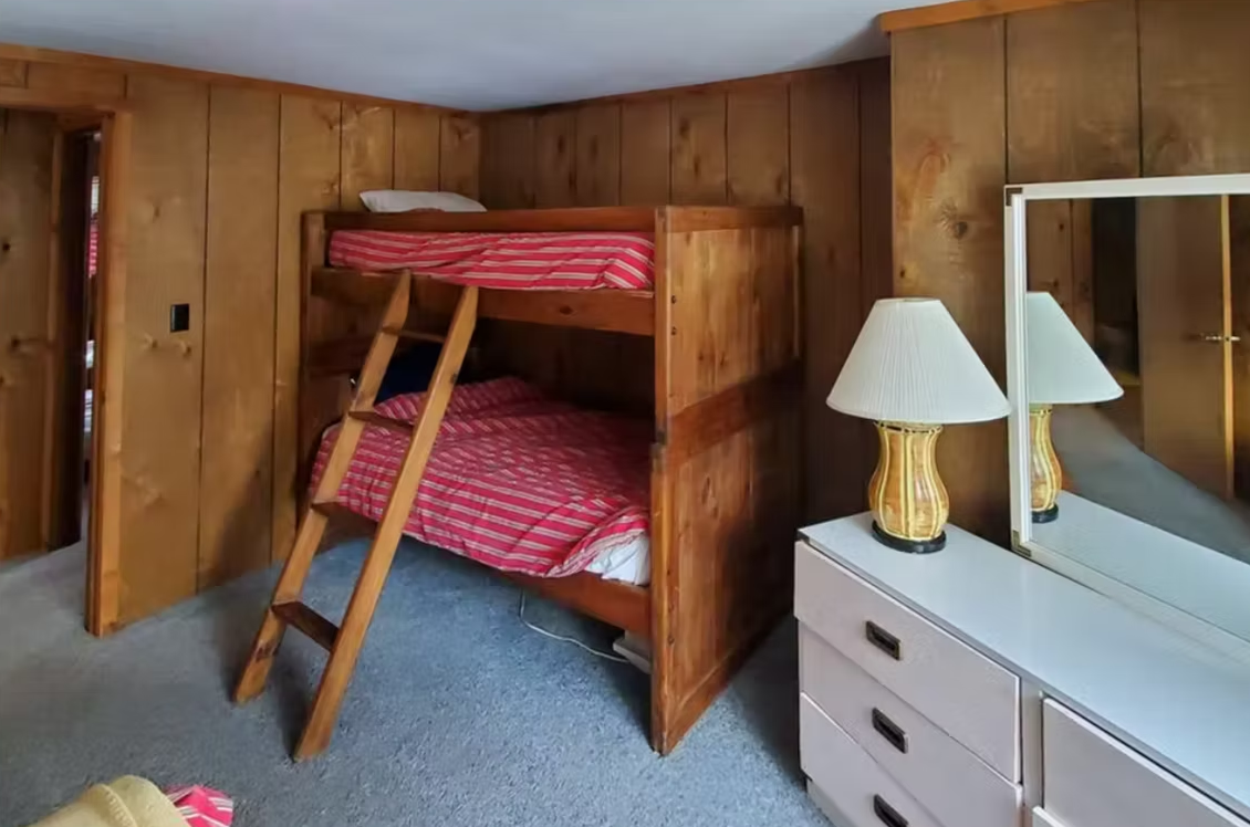 Wood-paneled bedroom with a wooden bunk bed, dresser, and lamp on a carpeted floor.