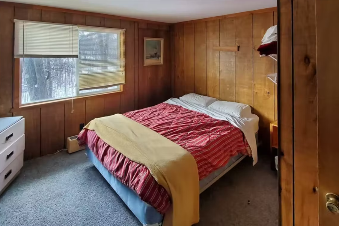 Cozy wood-paneled bedroom with a bed, red quilt, and window blinds.
