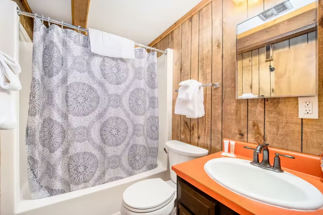 Small bathroom with wood paneling, orange sink counter, toilet, and shower with patterned curtain.