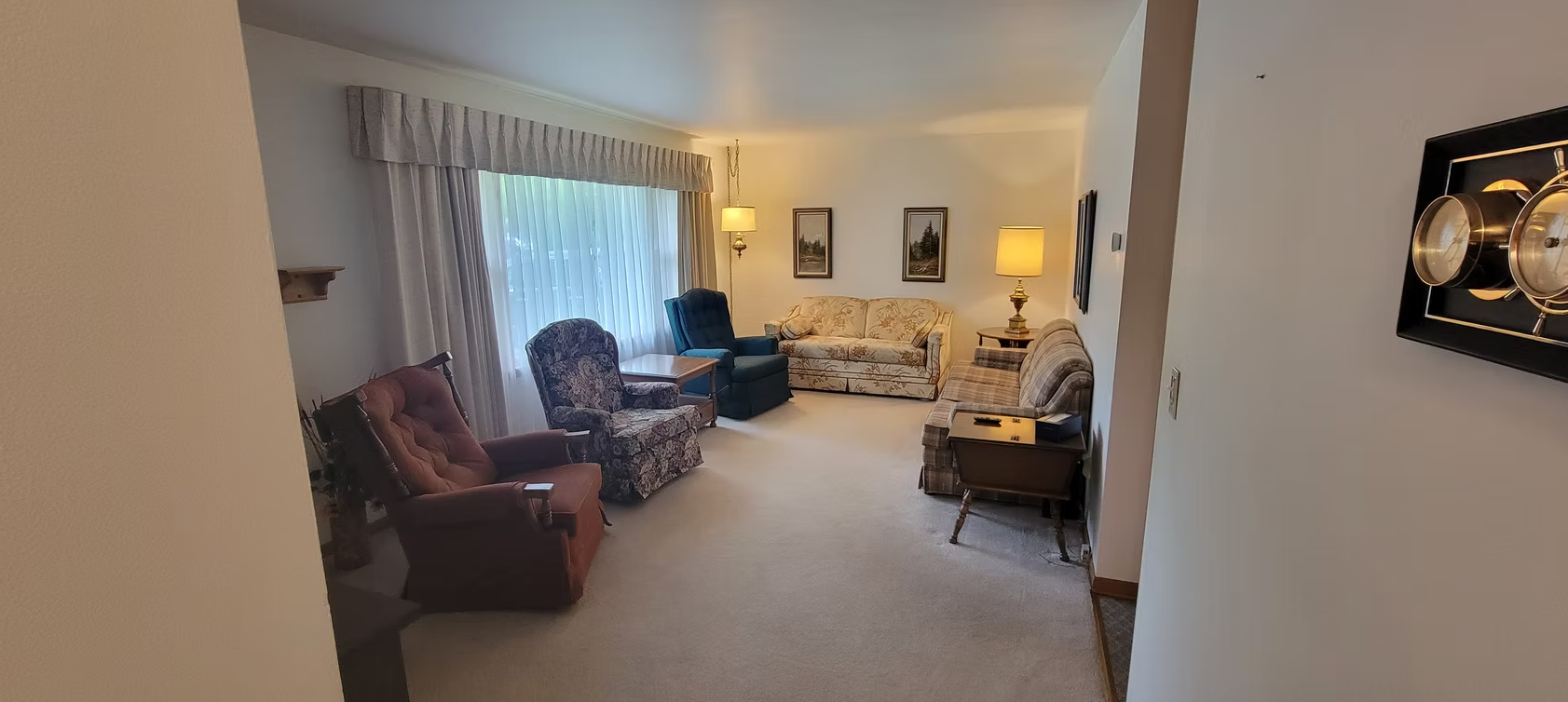 Cozy living room with chairs, sofa, lamp, framed art, and a large window with curtains.