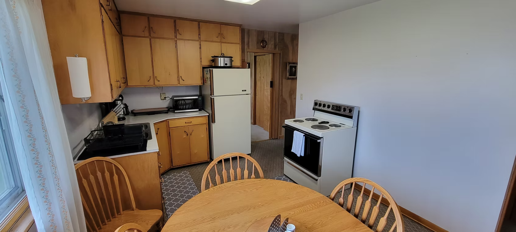 Small kitchen with wooden cabinets, white stove and fridge, and a dining table in the foreground.