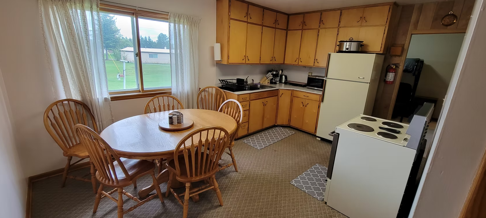 Small kitchen with wooden cabinets, round table and chairs, white stove and fridge, and a large window with sheer curtains.
