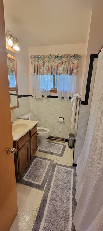 Small bathroom with sink vanity, toilet, window with floral curtain, and gray rugs