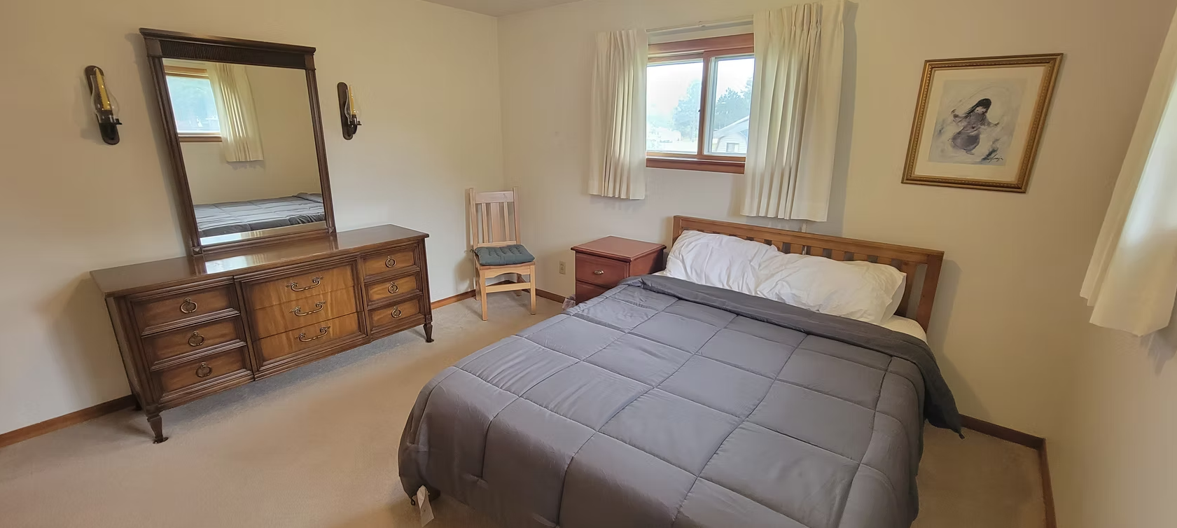 Bright bedroom with a bed, dresser, mirror, chair, and windows in a beige room