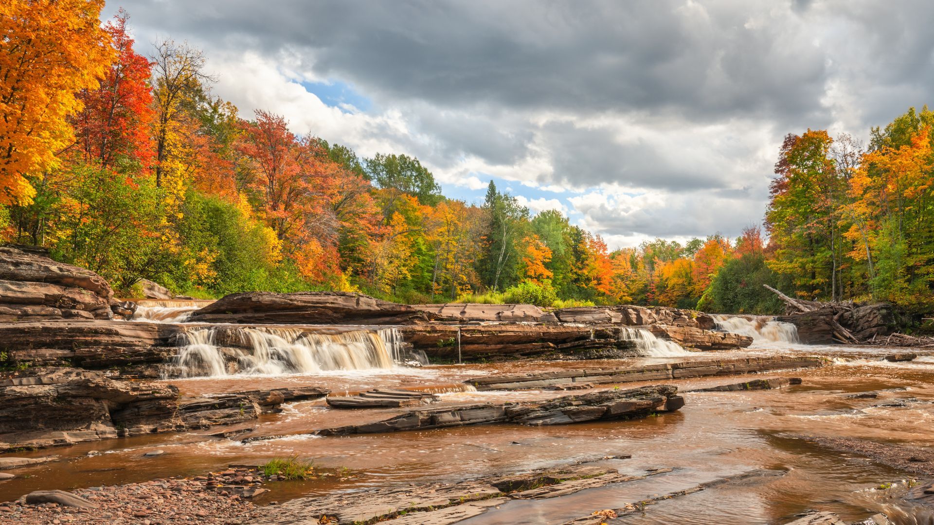 Autumn forest waterfall flowing over layered rocks under cloudy sky