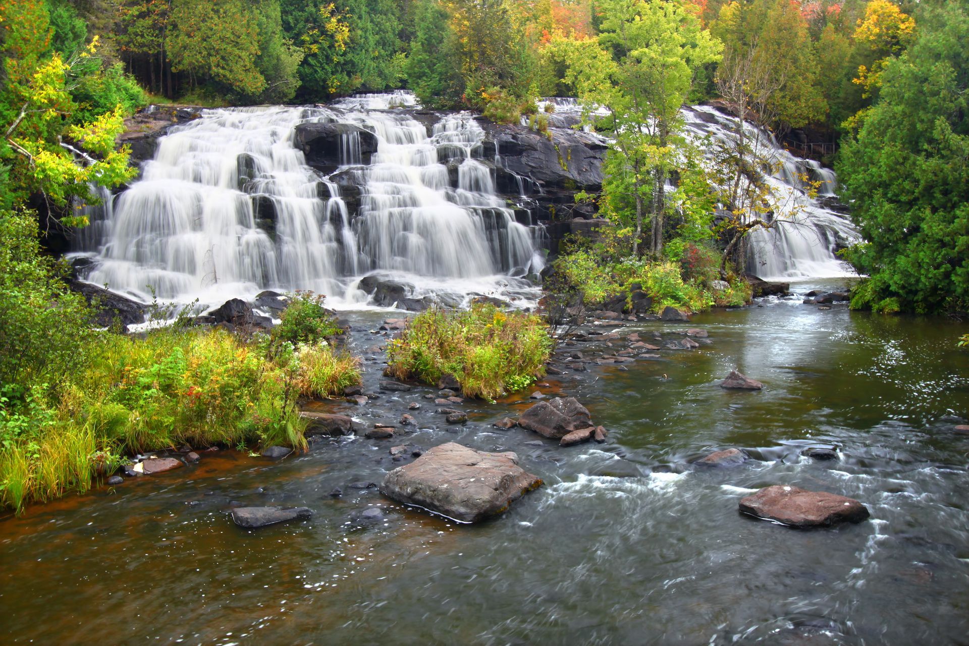 Waterfall cascading over rocks into a river, surrounded by autumn trees and greenery.