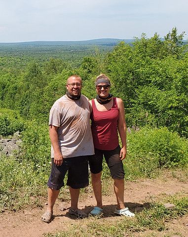 Two people standing on a dirt path with lush green hills in the background, smiling at the camera.