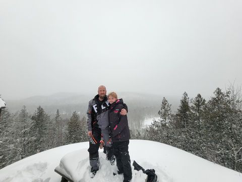 Two people standing on a snowy rock, with snow-covered trees and a foggy mountain backdrop.