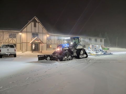 Snowplow clearing snow in front of a lit building at night, with snow-covered ground and parked vehicles.