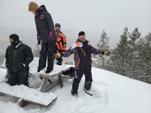 People standing in deep snow near a wooden bench on a snowy hillside with trees in the background