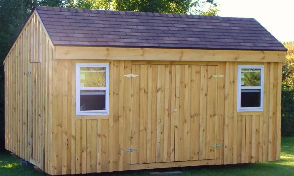 Wooden shed with brown roof and two white-framed windows, set in a grassy yard.