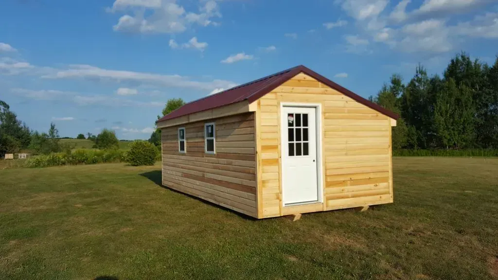Small wooden shed with white door and windows, brown roof, in a grassy field under a blue sky.