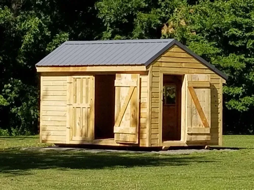 Wooden shed with gray metal roof and open doors, set in grassy area with trees.